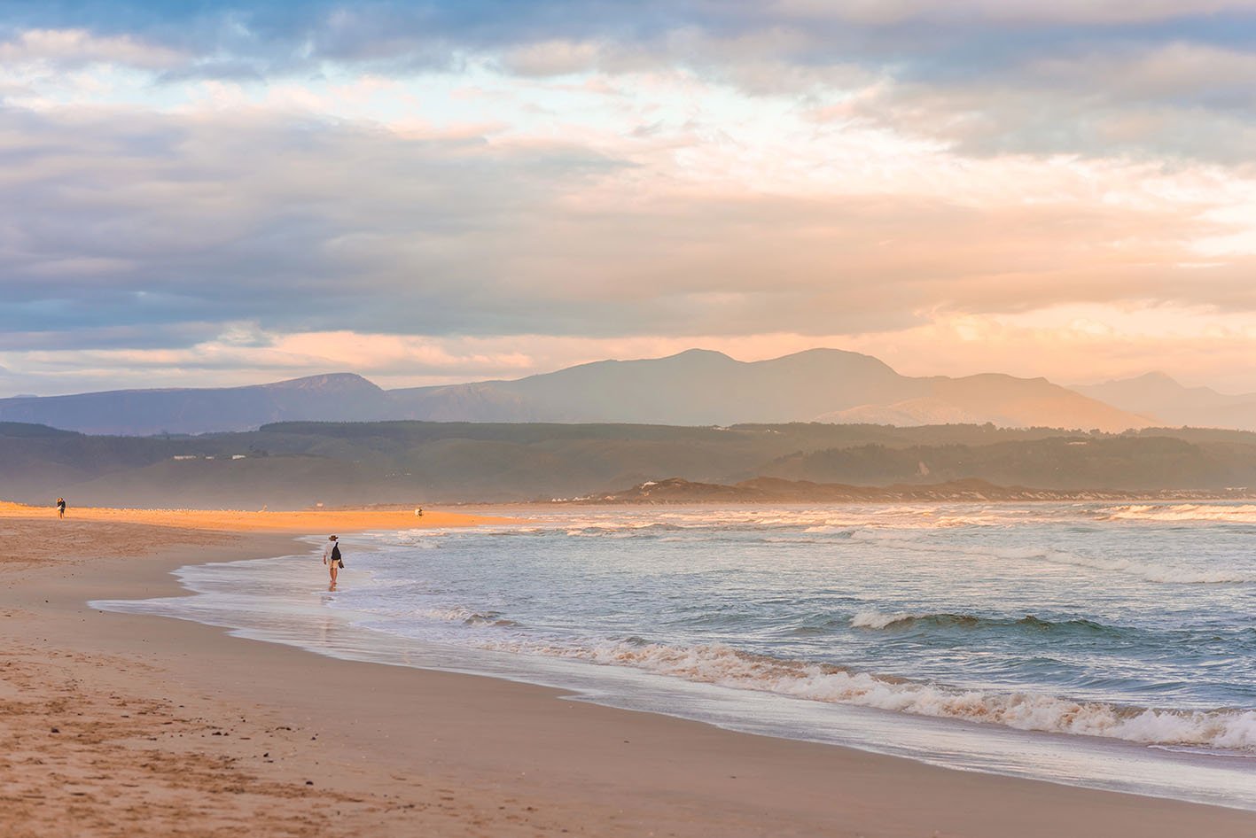 A streak of golden evening light falling on the Plettenberg Bay beach at sunset, with mountains in the distance and people walking on the beach. Garden Route, Western Cape, South Africa